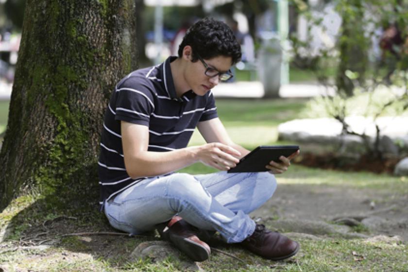 Un joven consulta una tableta digital sentado bajo un árbol