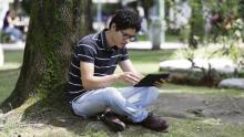 Un joven consulta una tableta digital sentado bajo un árbol